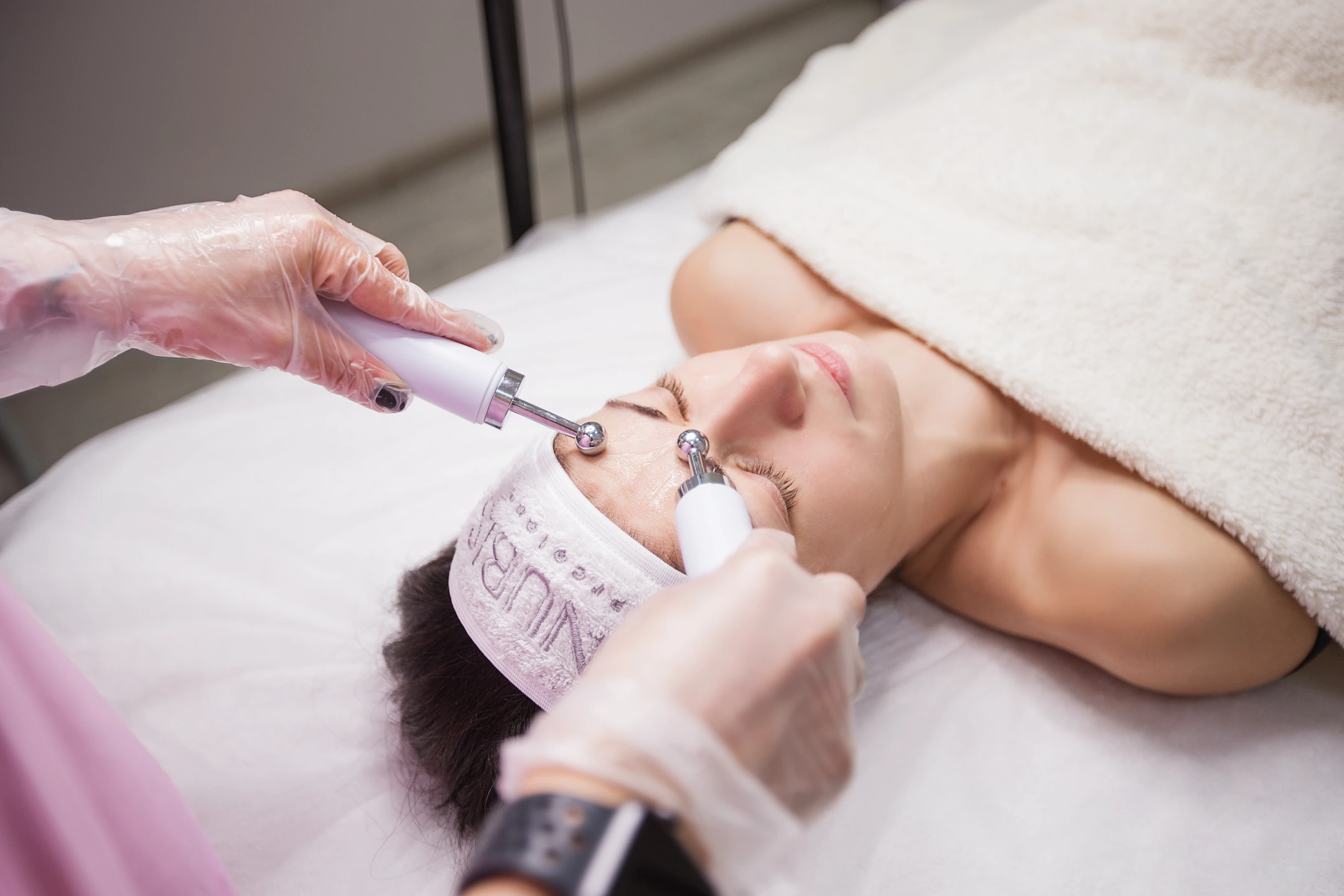 Gloved hands holding two rounded facial instruments against a client's forehead.