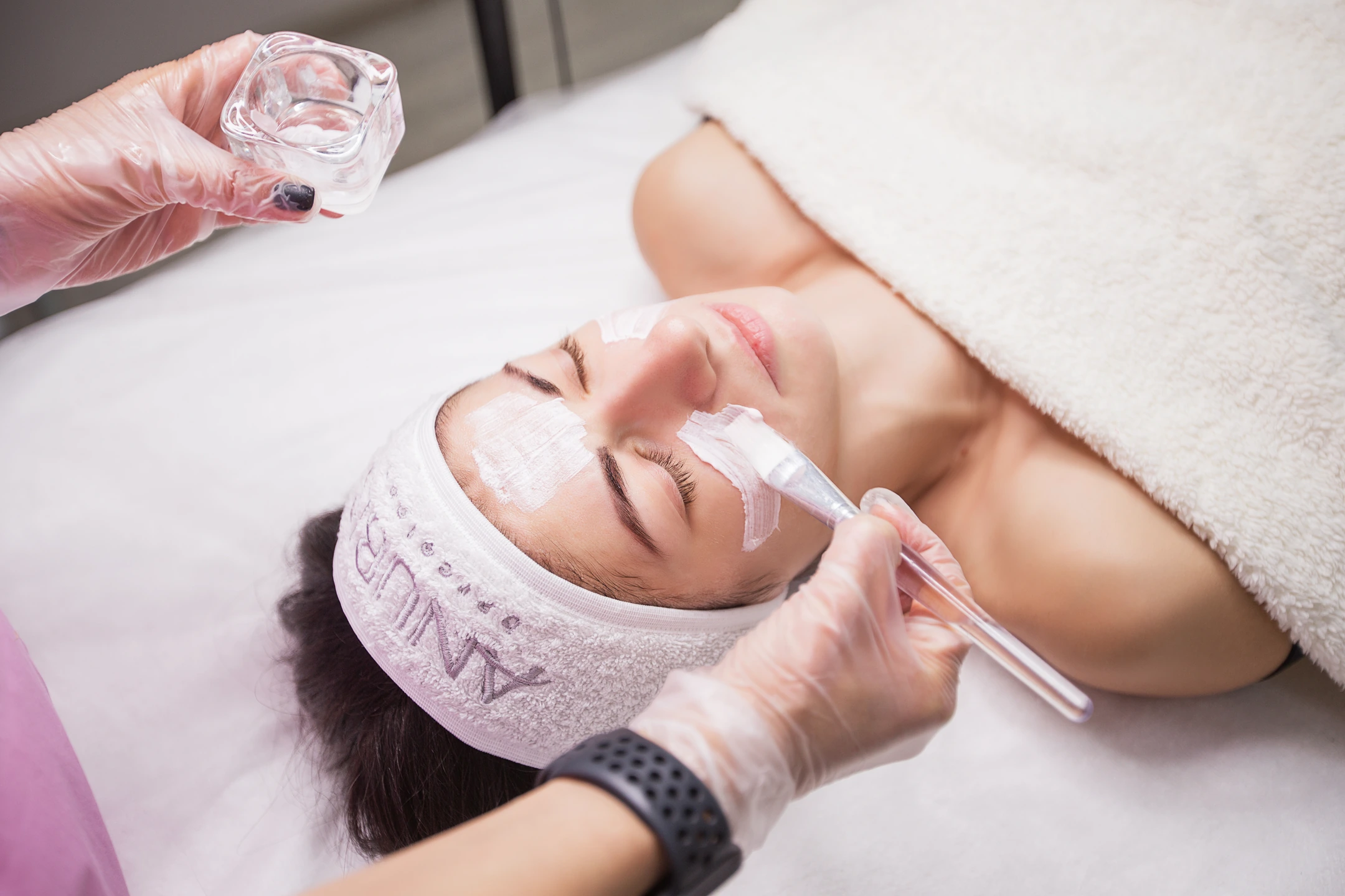 A brush applies a white facial mask during a treatment.
